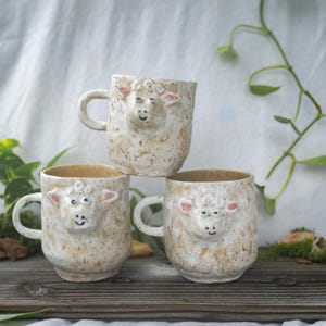 May include: Three ceramic mugs with a speckled beige finish, each featuring a sculpted, three-dimensional sheep face on the front. The mugs have a rounded handle on the side and a textured, earthy appearance with pink accents on the sheep ears.