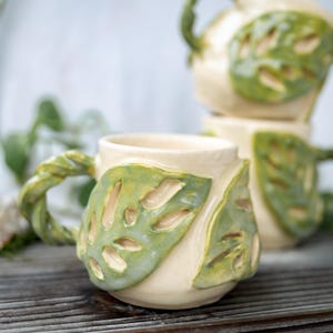 May include: Two ceramic mugs with a cream-colored base and raised, green, textured monstera leaf designs on the exterior. Each mug features a twisted, vine-like handle and a glossy finish, showcasing organic shapes and botanical-inspired details.
