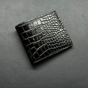 May include: A black bi-fold wallet with a crocodile skin texture. The wallet is made of a shiny, dark material and has a classic design. The wallet is closed and appears to be empty.