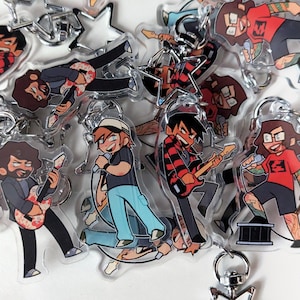 Puede incluir: Una colección de llaveros acrílicos transparentes con ilustraciones de dibujos animados de músicos. Los llaveros representan varios personajes tocando la guitarra y cantando en micrófonos, con colores vibrantes y diseños detallados. Cada llavero tiene un anillo de metal plateado.