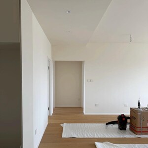 Op de afbeelding: Interieur van een nieuw gebouwde ruimte met witte muren, lichte houten vloeren en een verlaagd plafond. Een stofzuiger en een kartonnen doos zijn zichtbaar, samen met beschermende vloerbedekkingen. De ruimte omvat deuropeningen en een gang.