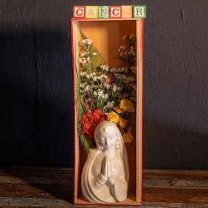 May include: A white ceramic statue of a praying figure is inside a wooden box, surrounded by flowers in shades of white, yellow, and red. The word "CANCER" is spelled out with colorful wooden blocks above the box. The box has a red border.