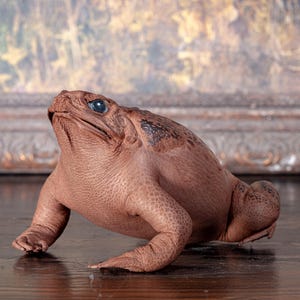 May include: A detailed, brown frog figurine with textured skin and blue eyes. The frog is posed in a forward-leaning position, with its front legs extended. The background is a blurred painting.