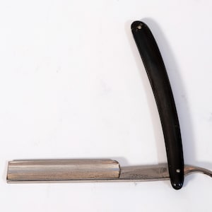 May include: A straight razor with a black handle and a silver blade. The blade is open, revealing its sharp edge. The razor is designed for shaving and grooming.