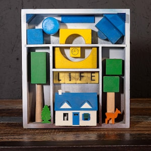 May include: A wooden display box filled with colorful wooden blocks. The blocks include a blue house, green trees, a yellow arch with a clock, and blocks spelling "LIFE". A small orange deer is also included.