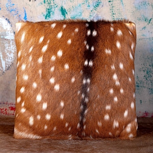 May include: A square decorative pillow with a brown and white spotted pattern, resembling deer hide. The pillow has a dark brown stripe down the center and is likely made of a soft, textured material. The pillow is a home decor item.