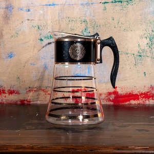 Puede incluir: Jarra de vidrio vintage con detalles en negro y dorado. La jarra presenta rayas horizontales doradas y una tapa negra con un emblema dorado decorativo. El asa es negra y curvada. Una pieza clásica para servir bebidas.