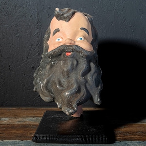 May include: A vintage ceramic head of a bearded man with blue eyes and a red mouth. The head is mounted on a black wooden base. The beard is dark gray and the face is painted with flesh tones.