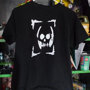 Può includere: T-shirt nero con un teschio bianco racchiuso in una cornice rettangolare. La maglietta è appesa a una gruccia bianca.