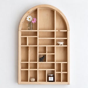 May include: A light-coloured wooden wall shelf with an arched top and multiple rectangular compartments of varying sizes. The shelf contains a small glass vase with two flowers, a white elephant figurine, a black elephant figurine, a small stone, and a jar.