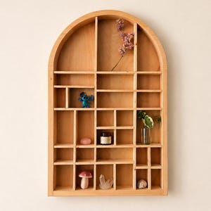 May include: Wooden arched display shelf with multiple compartments. The shelf is filled with small decorative items, including a blue figurine, a pink stone, a small jar, a glass vase with a plant, a mushroom figurine, and a crystal.