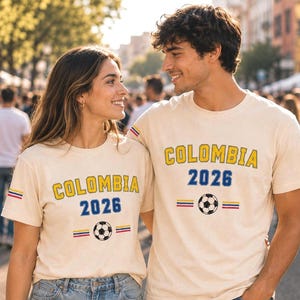 Op de afbeelding: Twee cr&egrave;mekleurige T-shirts met korte mouwen voorzien van de tekst COLOMBIA in het geel, het getal 2026 in het blauw, een voetbalafbeelding en kleine horizontale strepen in geel, blauw en rood op de mouwen en de borst.