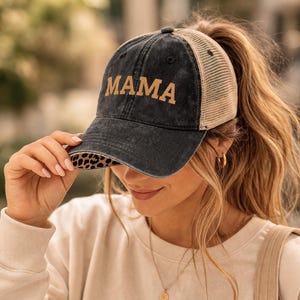 May include: A black and tan trucker hat with the word "MAMA" in gold lettering. The hat has a leopard print brim and a mesh back. The person is holding a coffee cup.