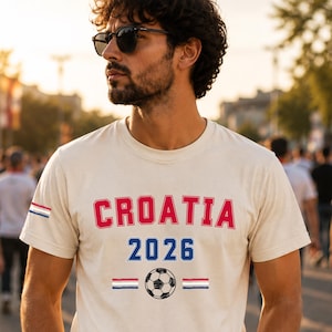 Puede incluir: Una camiseta de manga corta de color crema con el texto rojo CROATIA sobre el texto azul 2026. Debajo del texto hay un gr&aacute;fico de un bal&oacute;n de f&uacute;tbol en blanco y negro flanqueado por dos peque&ntilde;as rayas horizontales en rojo, blanco y azul.