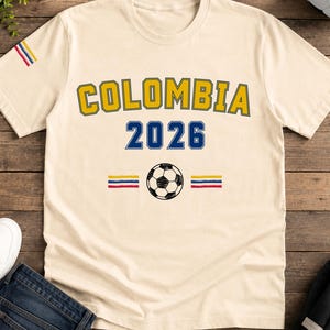 Op de afbeelding: Een cr&egrave;mekleurig t-shirt met korte mouwen met de tekst COLOMBIA in het geel met blauwe omlijning, het getal 2026 in het blauw, een zwart-witte voetbalafbeelding en horizontale gele, blauwe en rode strepen op de borst en de mouw.