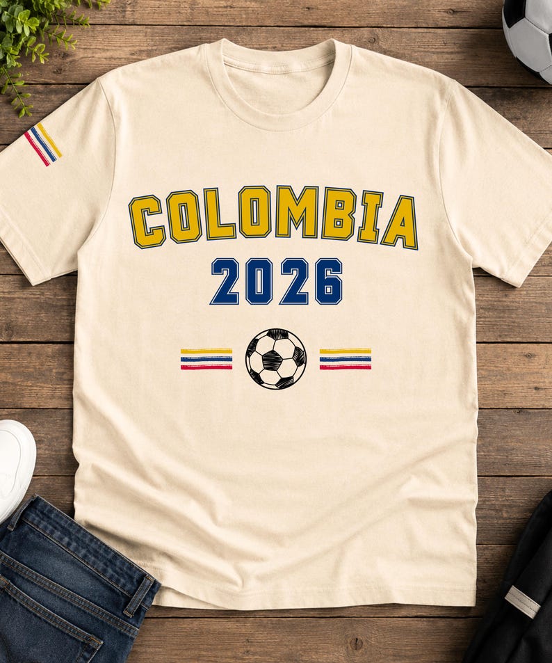 Op de afbeelding: Een cr&egrave;mekleurig t-shirt met korte mouwen met de tekst COLOMBIA in het geel met blauwe omlijning, het getal 2026 in het blauw, een zwart-witte voetbalafbeelding en horizontale gele, blauwe en rode strepen op de borst en de mouw.