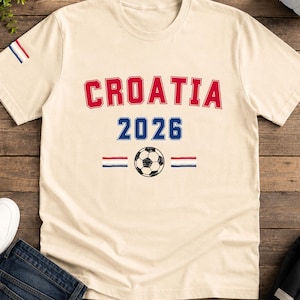 Puede incluir: Una camiseta de manga corta color crema con el texto rojo CROATIA arqueado sobre el texto azul 2026. Debajo del a&ntilde;o hay un gr&aacute;fico de un bal&oacute;n de f&uacute;tbol en blanco y negro flanqueado por dos peque&ntilde;os iconos de banderas a rayas rojas, blancas y azules.