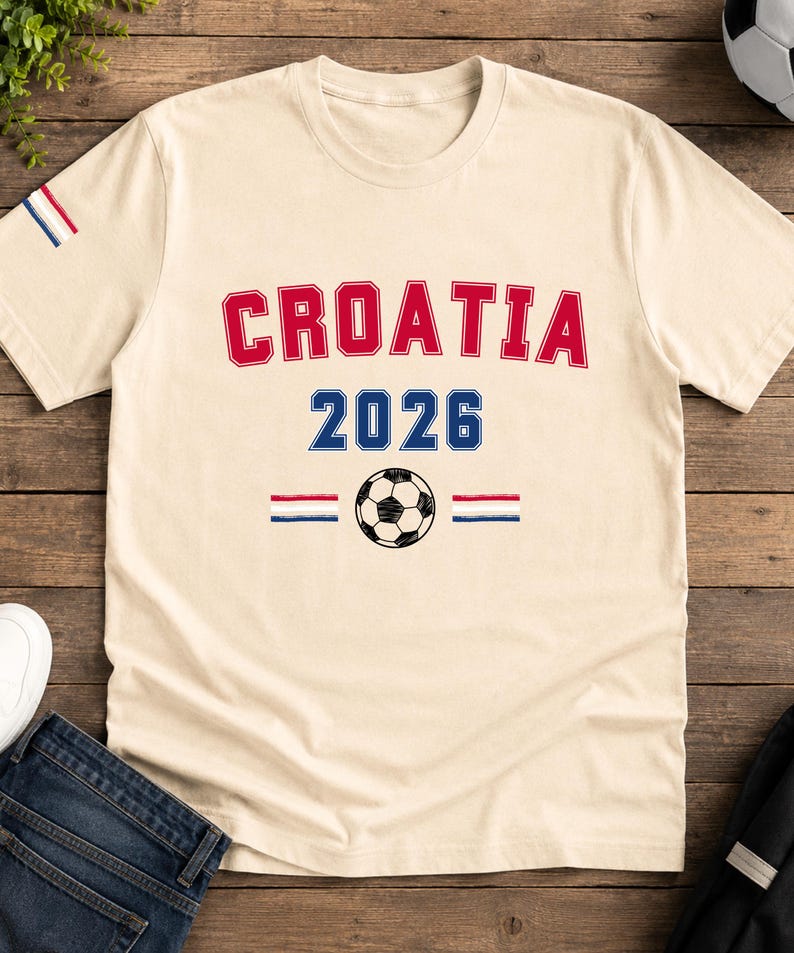 Puede incluir: Una camiseta de manga corta color crema con el texto rojo CROATIA arqueado sobre el texto azul 2026. Debajo del a&ntilde;o hay un gr&aacute;fico de un bal&oacute;n de f&uacute;tbol en blanco y negro flanqueado por dos peque&ntilde;os iconos de banderas a rayas rojas, blancas y azules.
