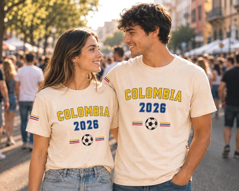 Op de afbeelding: Twee cr&egrave;mekleurige T-shirts met korte mouwen voorzien van de tekst COLOMBIA in het geel, het getal 2026 in het blauw, een voetbalafbeelding en kleine horizontale strepen in geel, blauw en rood op de mouwen en de borst.