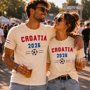 Puede incluir: Dos camisetas de manga corta color crema con el texto rojo CROATIA, el texto azul 2026, un gr&aacute;fico de bal&oacute;n de f&uacute;tbol en blanco y negro, y peque&ntilde;as rayas horizontales rojas, blancas y azules en la parte frontal.