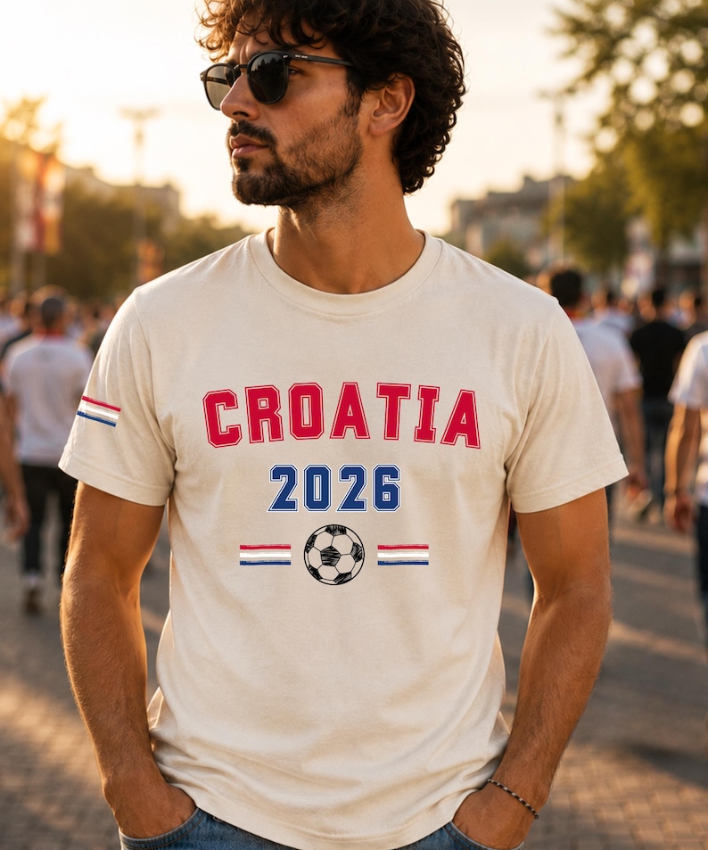 Puede incluir: Una camiseta de manga corta de color crema con el texto rojo CROATIA sobre el texto azul 2026. Debajo del texto hay un gr&aacute;fico de un bal&oacute;n de f&uacute;tbol en blanco y negro flanqueado por dos peque&ntilde;as rayas horizontales en rojo, blanco y azul.
