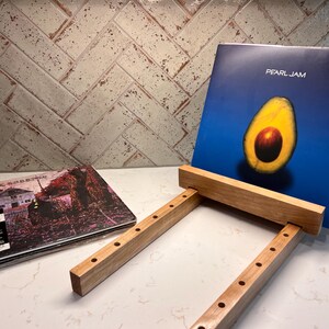 May include: A light-colored wooden vinyl record display stand with a series of circular holes along the side rails. A Pearl Jam album with an avocado graphic is resting on the stand, and a Black Sabbath vinyl record is lying flat nearby.