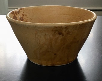 May include: A large, off-white ceramic bowl with a slightly flared rim. The bowl has a rustic, hand-thrown look with brown and tan streaks and spots throughout.