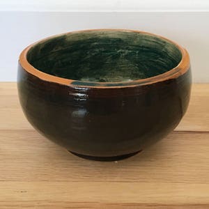 Op de afbeelding: Een handgemaakte keramische kom met een glanzende, donkerbruine buitenkant en een groene binnenkant. De rand is lichtbruin van kleur. De kom is rond en staat op een kleine, cirkelvormige basis. De kom staat op een houten oppervlak.