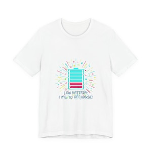 Puede incluir: Camiseta blanca con un diseño gráfico colorido de un icono de batería baja y el texto "LOW BATTERY: TIME TO RECHARGE!". El diseño incluye un contorno de batería turquesa con relleno rojo, rodeado de formas coloridas.