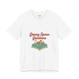 May include: White t-shirt featuring the phrase "Greasy Spoon Greatness" in a retro font. Below the text is a colorful illustration of a diner table with a burger, fries, milkshake, and coffee. The shirt has a crew neck and short sleeves.