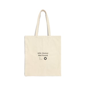 May include: A cream-coloured tote bag featuring the text "404: Humor Not Found" in black, along with a gear and line graphic. The bag has two handles and is made of a canvas-like material.