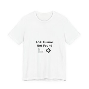 Puede incluir: Camiseta blanca con el texto negro "404: Humor Not Found", junto con un circuito y un engranaje. La camiseta de manga corta está hecha de un material suave.