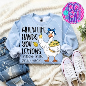 May include: Light blue sweatshirt with a graphic of a goose wearing a blue bow and holding lemons. Text on the shirt reads "WHEN LIFE HANDS YOU LEMONS throw that shit back!" The sweatshirt is paired with jeans and white sneakers.