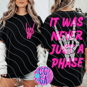 May include: Black t-shirt with a pink skeleton hand rock and roll graphic on the front. The back of the shirt features the text "IT WAS NEVER JUST A PHASE" in pink and gray. The shirt is a casual, unisex style.