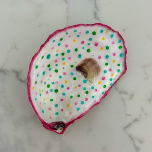 May include: Hand-painted oyster shell with a white interior, a pink rim, and colorful polka dots in green, blue, pink, and yellow. The shell features a natural, brown, shell-like accent in the center.