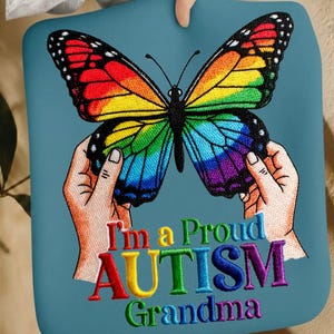 May include: A teal-colored cushion featuring a vibrant butterfly with rainbow-colored wings. The butterfly is held by two hands. The text "I'm a Proud AUTISM Grandma" is embroidered in colorful letters.