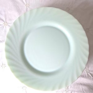 Puede incluir: Un plato llano redondo de color verde menta pálido con un centro liso y plano, y un borde ancho decorativo que presenta un sutil patrón radial en espiral que se extiende desde el círculo interior hasta el borde exterior ondulado y festoneado.