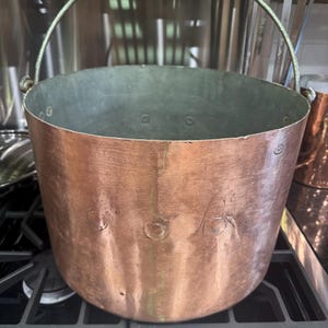 May include: A copper cooking pot with a riveted construction and a sturdy metal handle. The pot has a wide, open mouth and a slightly rounded base. The copper has a warm, reddish-brown hue, and the interior is a contrasting gray color.