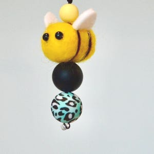 May include: A hanging ornament featuring a yellow and black felt bee with small white wings, suspended by a black cord. Below the bee are two spherical beads: one solid matte black bead and one turquoise bead with a black leopard print pattern.