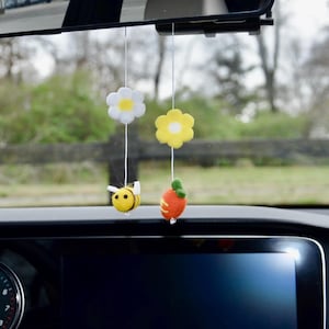 May include: Car hanging decorations featuring a white and yellow daisy, a yellow flower, a yellow and black bee, and an orange carrot with a green top. The ornaments are suspended from a car's rearview mirror.