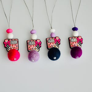 May include: Four colorful teacher-themed car air fresheners. Each has a felt ball in a different color: pink, purple, navy, and dark purple. They also have a book-shaped charm with the word "TEACHER" on it, along with a bow and flower design.