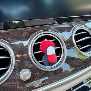 May include: A car air vent accessory shaped like a red, white, and blue pill, with a small red pom-pom hat and a dark blue pom-pom. The accessory is placed in a car's air vent, with a dark wood grain dashboard.
