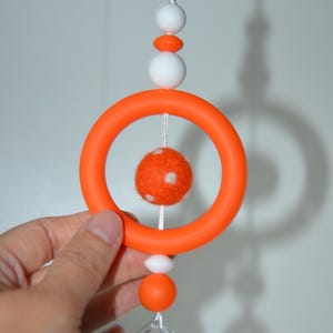 May include: A hanging mobile with an orange ring, white and orange beads, and a crystal pendant. Inside the ring hangs a small orange felt ball with white spots. The mobile is suspended by a white cord.