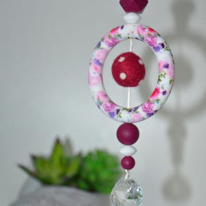 May include: A decorative hanging mobile with a floral-patterned ring, a red felt ball, and a crystal pendant. The mobile features burgundy and white beads, and a burgundy geometric bead at the top. The floral pattern includes pink and purple flowers.
