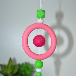 May include: A hanging mobile featuring a pink ring, a felt ball, green and white beads, and a green geometric shape. The mobile is suspended by a white string, creating a playful and colorful decorative item.