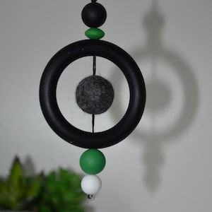 May include: A contemporary hanging mobile with a black ring and a grey felt ball at its core. The mobile is decorated with black, green, and white spherical beads. The design is suspended by a black string, creating a minimalist, decorative piece.