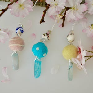 May include: Three handmade wind chimes with felt spheres in pink, blue, and yellow, each adorned with beads and a crystal pendant. The chimes hang near a branch with pink cherry blossoms.