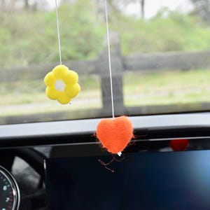 May include: Two car air fresheners suspended from a car's rearview mirror. One is a yellow flower with a white centre, and the other is an orange heart. The background shows a blurred green landscape.