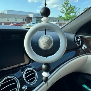May include: A car air freshener with a white ring and a grey felt ball at its centre, suspended by a black string. Black and white beads are above the ring. The accessory hangs in front of a car dashboard.