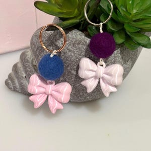 May include: Two keychains featuring metal rings, one with a dark blue felt ball and a light pink bow charm, and the other with a deep purple felt ball and a light pink bow charm. The charms are attached to the rings by small white cords.